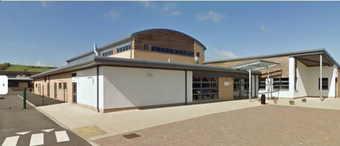 A school building with a curved roof. The building is tan and brown. A paved area is in front