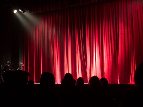 An audience waits in front of a red curtain and stage